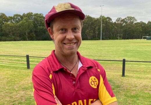 Jason Fergus stands on the Bulleen Park grounds. He is wearing full Yaraleen Cricket club uniform. he is smiling and his arms are crossed.